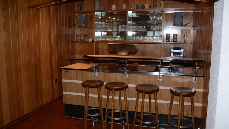 Eine gemütliche Bar mit Holzwänden und einer langen Theke. Vier Barhocker stehen davor, bereit für Gäste.