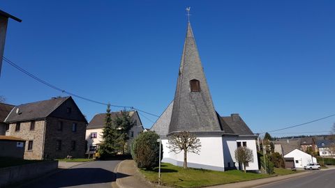 Eine Kirche mit einem spitzen Turm steht in einer ruhigen Straße. Im Hintergrund sind traditionelle Häuser und ein klarer blauer Himmel zu sehen.