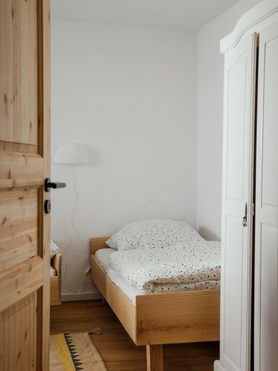 Ein einfaches Schlafzimmer mit zwei Betten und hellen Bettdecken. Der Raum hat einen Holztür und einen weißen Kleiderschrank.