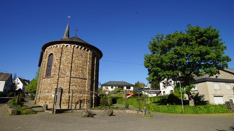 Eine historische Kirche mit einer runden Form aus Steinen. Im Hintergrund sind Häuser und ein großer Baum zu sehen, unter einem klaren blauen Himmel.