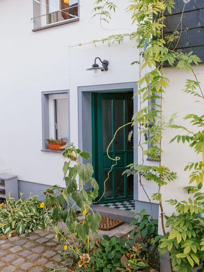 Eine einladende Haustür in einem Garten mit vielen Pflanzen. Der Eingang ist von grünen Ranken und bunten Blumen umgeben.