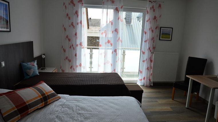 A bright room with two beds and a table. Large windows with white curtains let in plenty of light.
