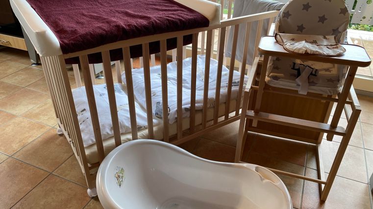 Ein Babybett mit einer weißen Matratze und rotem Bezug steht in einem Raum. Neben dem Bett sind ein Hochstuhl und eine Babybadewanne zu sehen.