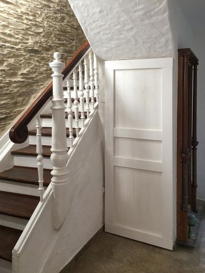 Eine helle Treppe mit weißem Geländer führt nach oben. Neben der Treppe steht eine weiße Tür, die in einen schlichten Raum führt.