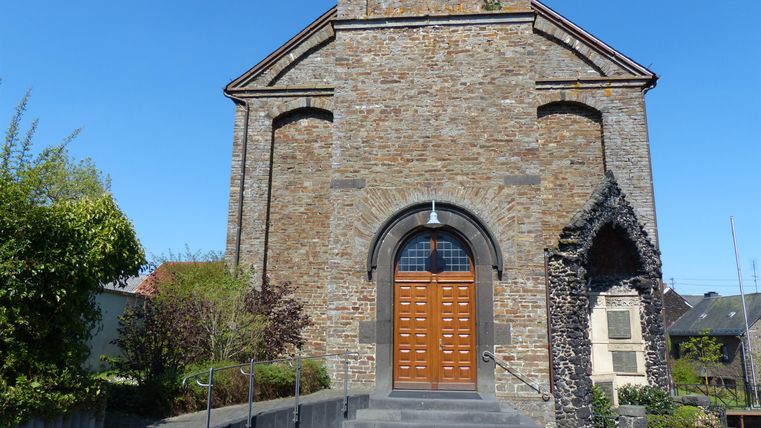 Eine steinerne Kirche mit einem großen Holztor. Der Himmel ist klar und blau, umgeben von grünen Bäumen.