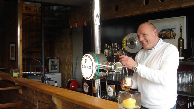 Ein fröhlicher Barkeeper zapft Bier an der Theke. Im Hintergrund sind Getränke und eine gemütliche Atmosphäre zu erkennen.