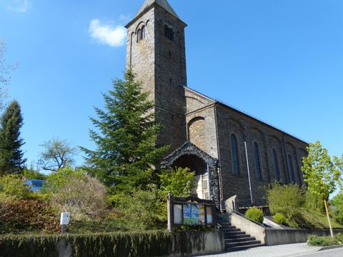 Eine Kirche mit einem hohen Turm und steiniger Fassade steht vor einem klaren blauen Himmel. Grüne Bäume und Sträucher umgeben das Gebäude.