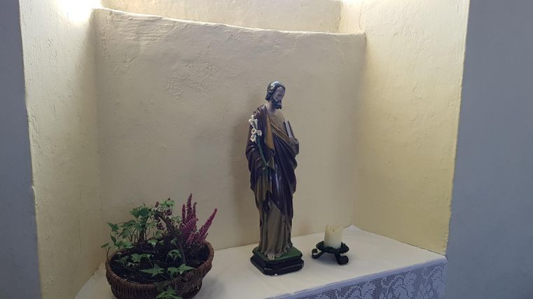 Eine kleine Nische mit einer Statue und einer Kerze. Daneben steht ein Pflanzentopf mit grünen Pflanzen und lila Blüten.