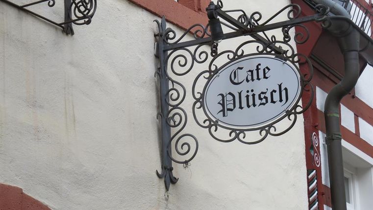 Ein dekoratives Schild eines Cafés mit dem Namen "Café Plüsch". Das Schild hängt an einer hellen Wand in einer charmanten Altstadt.