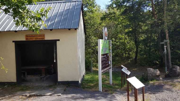 A small building with a metal roof and a wooden sign. Next to the building, there are information signs near a forest.