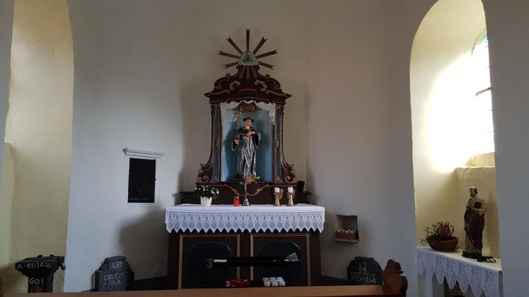 Ein Innenraum einer kleinen Kirche mit einem Altar und einer Statue. Der Altar ist mit einem weißen Tuch und Kerzen dekoriert.