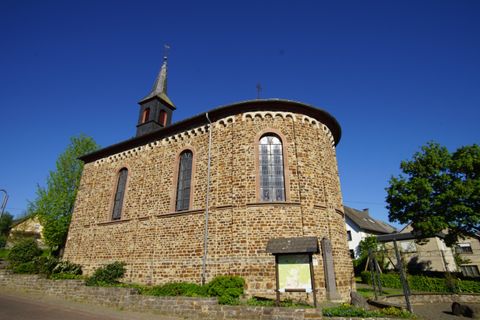 A historic church with a round shape and a bell tower. The sky is clear and blue, surrounded by trees and a peaceful village.