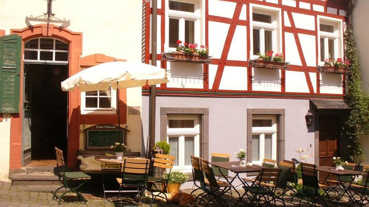 Ein gemütliches Café mit Tischen und Stühlen im Freien. Die Fassade ist traditionell mit Holzrahmen und blühenden Fensterkästen dekoriert.