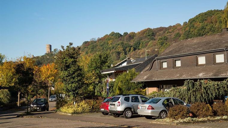 Ein ruhiger Parkplatz mit mehreren Autos und Bäumen in der Umgebung. Im Hintergrund sind Hügel und ein Aussichtsturm sichtbar.