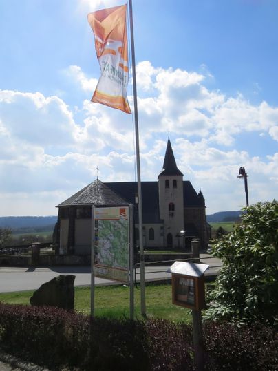 Eine kleine Kirche mit einem spitzen Turm steht unter einem blauen Himmel. Neben der Kirche befindet sich ein Wegweiser und eine Fahne.