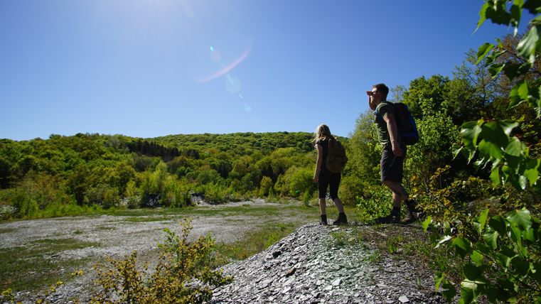 Zwei Wanderer stehen auf einem Schieferhügel und blicken in die Ferne, umgeben von grüner Vegetation und blauem Himmel.