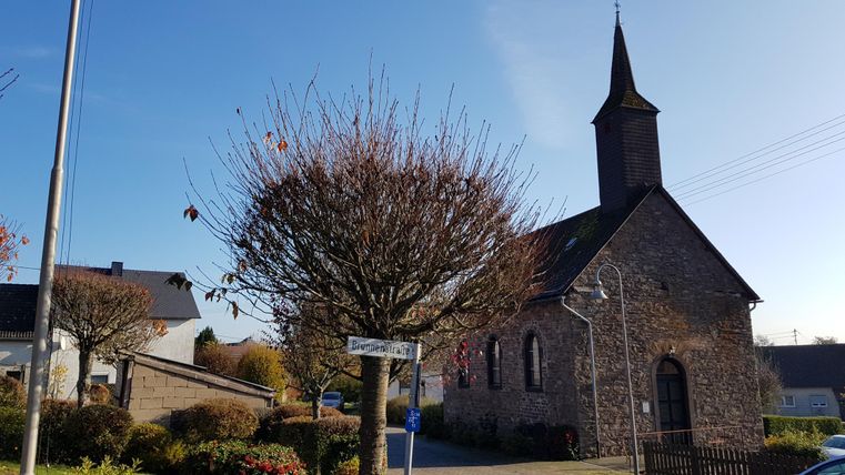 Eine kleine Kirche mit einem hohen Turm steht in einer ruhigen Straße. Rund um die Kirche sind viele Bäume und blühende Pflanzen.