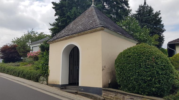 Eine kleine Kapelle mit einem schrägen Dach steht am Straßenrand. Grüne Sträucher und Bäume umgeben das Gebäude.