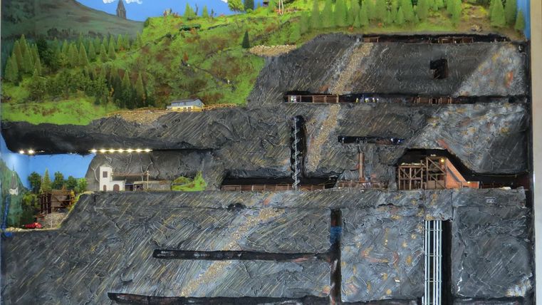 Ein Modell einer Bergbaulandschaft mit verschiedenen Schichten und Strukturen. Die Darstellung zeigt Bäume, Gebäude und Bergwerkseingänge.