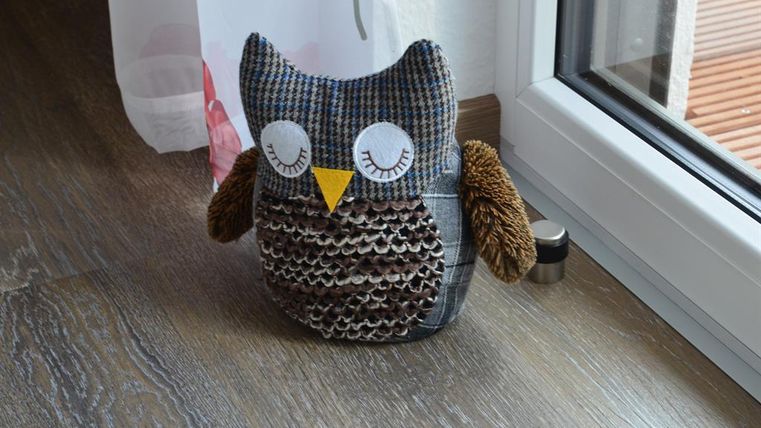 A cute fabric owl with large, white eyes and a pointed beak. It stands on a wooden floor next to a window.
