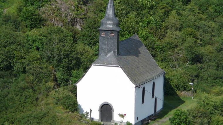 Eine kleine, weiße Kirche mit einem spitzen Turm steht umgeben von grüner Vegetation. Die ruhige Lage bietet eine idyllische Atmosphäre.