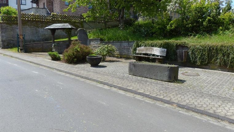 Eine ruhige Straßenszene mit einer Bank und einem überdachten Sitzbereich. Im Hintergrund sind grüne Büsche und ein steinernes Mauerwerk zu sehen.