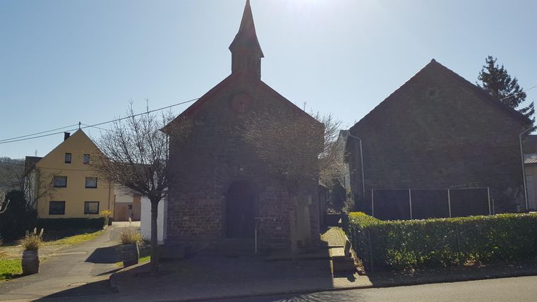 Eine kleine, steinerne Kapelle mit einem spitzen Turm steht neben einem älteren Gebäude. Im Hintergrund sind Häuser und ein blauer Himmel zu sehen.