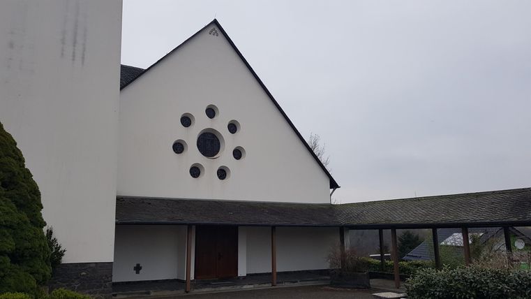 A modern church building with a white facade and a round window. In the background, a gray sky can be seen.