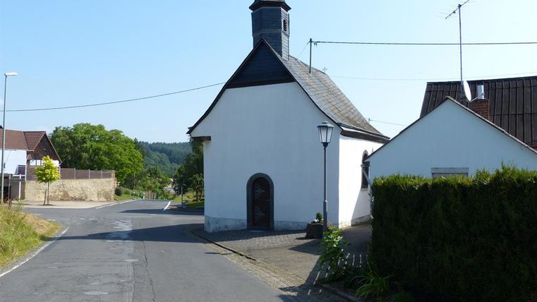 Eine kleine, weiße Kirche steht an einer Kreuzung. Im Hintergrund sind Bäume und Häuser zu sehen.