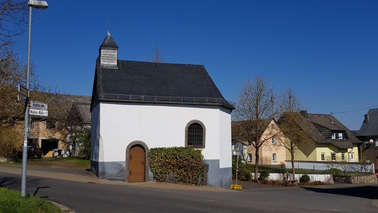 Eine kleine weiße Kapelle mit einem schwarzen Dach steht an einer ruhigen Straße. Im Hintergrund sind traditionelle Häuser und blauer Himmel zu sehen.