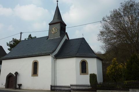 A small church with a pointed tower and a clock. It has white walls and is surrounded by trees.