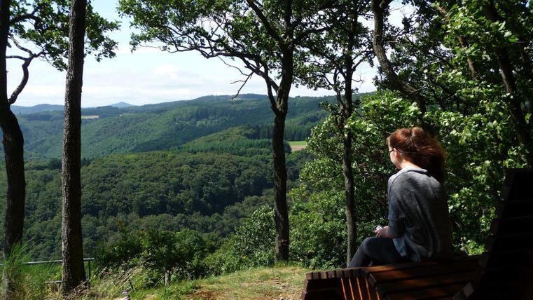 Eine Person sitzt auf einer Bank und schaut auf die bewaldete Hügellandschaft. Der Himmel ist klar und die Natur wirkt friedlich.