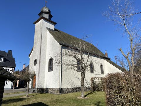 Eine charmante Kirche mit einem schlichten, weißen Äußeren und einem kleinen Glockenturm. Im Vordergrund sind kahle Bäume und ein gepflegter Rasen zu sehen.