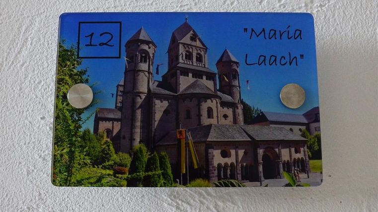 An image of the Maria Laach Abbey with several towers and a friendly garden landscape. The facade is made of gray stone and the architecture is historic.