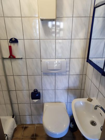 Ein einfaches Badezimmer mit weißen Fliesen. Es enthält eine Toilette, ein Waschbecken und einige Hygienemittel.