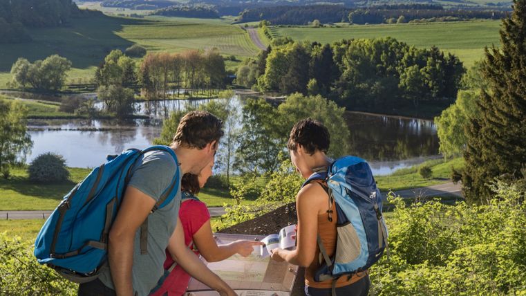 Three people carrying backpacks are looking at an information sign in front of a landscape with a maar and hills in the background.