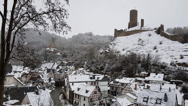 Ein malerisches Dorf mit schneebedeckten Dächern und historischen Gebäuden. Im Hintergrund erhebt sich eine alte Burg auf einem Hügel.