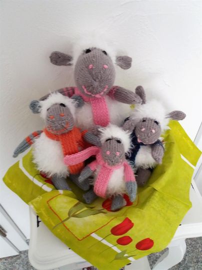 A group of four hand-knitted sheep sits in a colorful bag. The sheep have different colors and accessories.