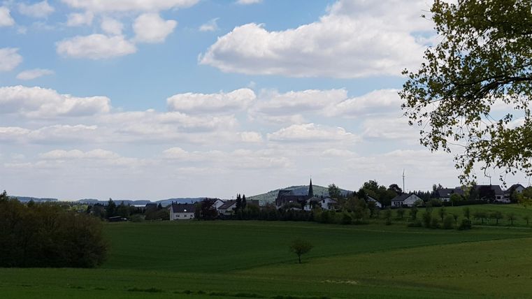 A picturesque landscape with gentle hills and expansive green fields. In the background, houses and a church are visible under a cloudy sky.