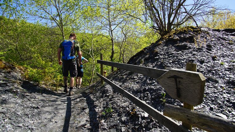 Two hikers on a narrow path in a slate landscape, surrounded by green trees and blue sky.