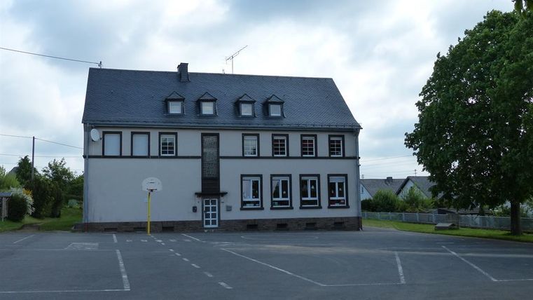 Ein großes, zweigeschossiges Gebäude mit einem Satteldach und mehreren Fenstern. Es steht auf einem Parkplatz mit einem Basketballkorb im Vordergrund.
