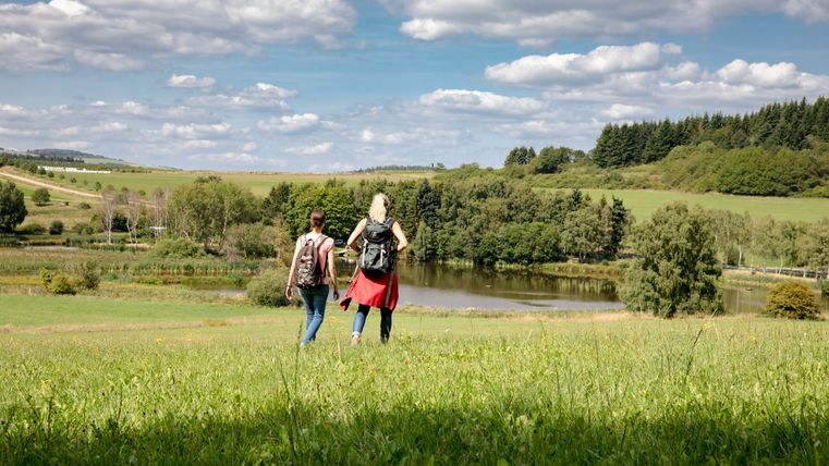 Zwei Personen wandern auf einer Wiese mit Blick auf einen See und bewaldete Hügel im Hintergrund.