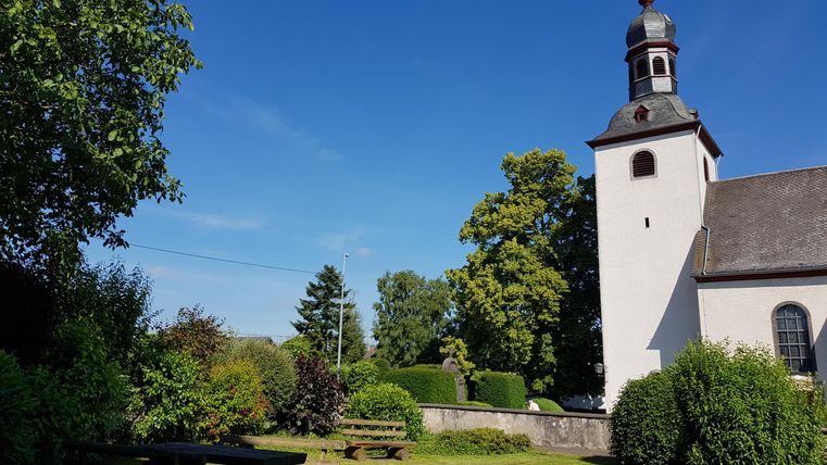 Eine sonnige Landschaft mit einer Kirche und grünem Rasen. Im Vordergrund sind Holztische und Bänke zu sehen.