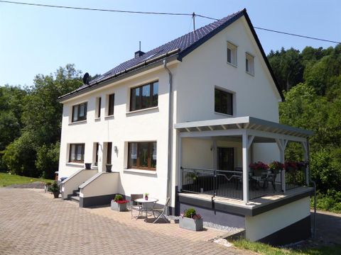 Ein modernes Haus mit einem großen Balkon und einer Terrasse. Umgeben von Grün und einem gepflasterten Gartenbereich.