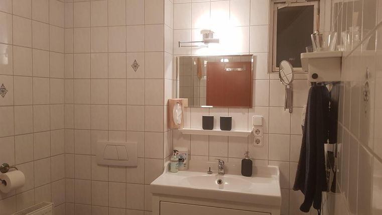 A small bathroom with white tiles and a sink. There is a mirror, a toilet, and a small window.