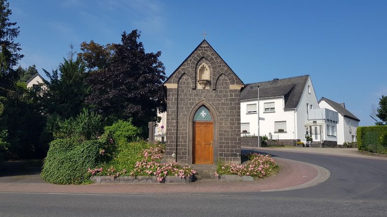 Eine kleine Kapelle aus dunklem Stein steht an einer Straßenkurve. Grüne Pflanzen und Blumen umrahmen den Eingang.