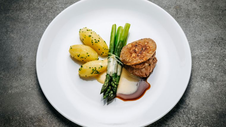 Ein Teller mit zartem Fleisch, grünem Spargel und Salzkartoffeln. Das Gericht ist kunstvoll angerichtet und mit einer Sauce verfeinert.