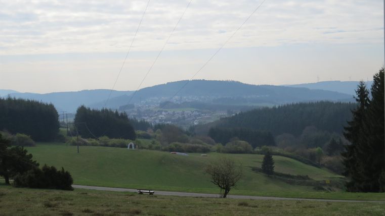 Eine weite Landschaft mit sanften Hügeln und Bäumen. Im Hintergrund sind Wolken am Himmel zu sehen.