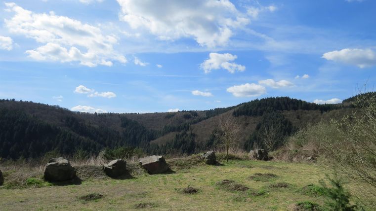 Eine weite Landschaft mit sanften Hügeln und klar blauem Himmel. Im Vordergrund liegen einige große Steine auf einer Wiese.