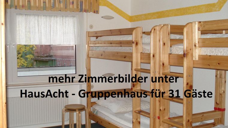 Ein einfaches Zimmer mit zwei Etagenbetten aus Holz und einem kleinen Tisch. Das Zimmer hat ein Fenster mit Vorhängen und einen neutralen Teppichboden.